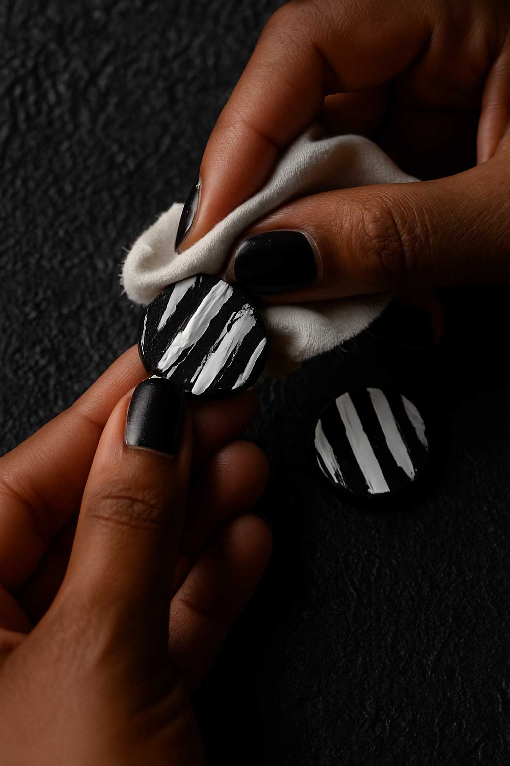 Paola Borde polissant une boucle d’oreille en liège noir et blanc à l’aide d’un chiffon doux, sur un fond texturé noir. Geste artisanal signé Paola Borde.
