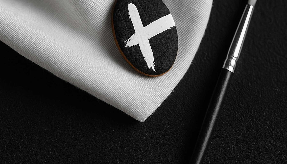 Boucle d’oreille en liège noir peinte à la main en blanc, posée sur un tissu blanc délicat, avec un pinceau fin à côté, sur fond noir texturé. Une composition sobre illustrant l’élégance du minimalisme signé Paola Borde
