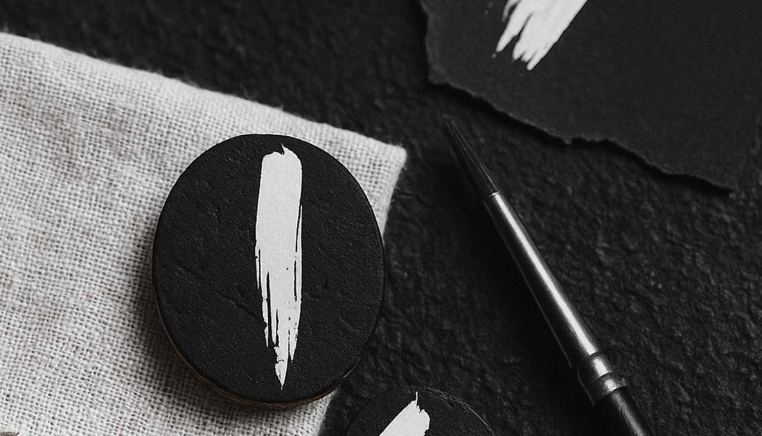 Bijoux en liège noir peints en blanc, posés avec pinceau et papier, illustrant l’équilibre entre graphisme et artisanat